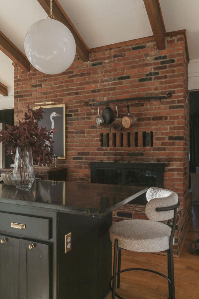 exposed brick fireplace in moody cottage kitchen with copper pans on hanging on wall and black cabinets
