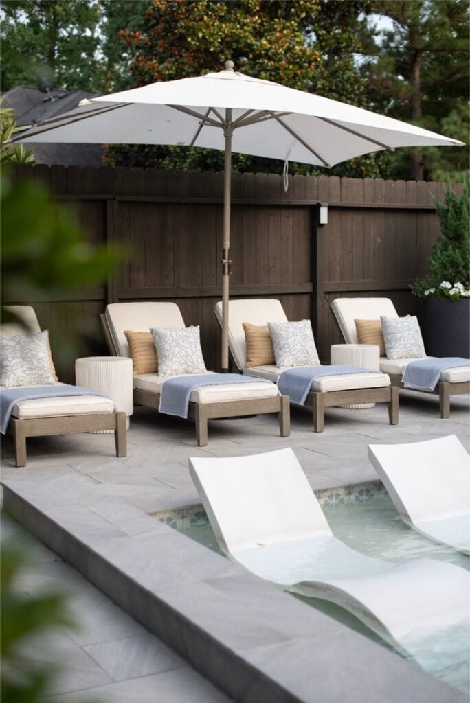 four chaise loungers by pool with one long rectangular umbrella in center